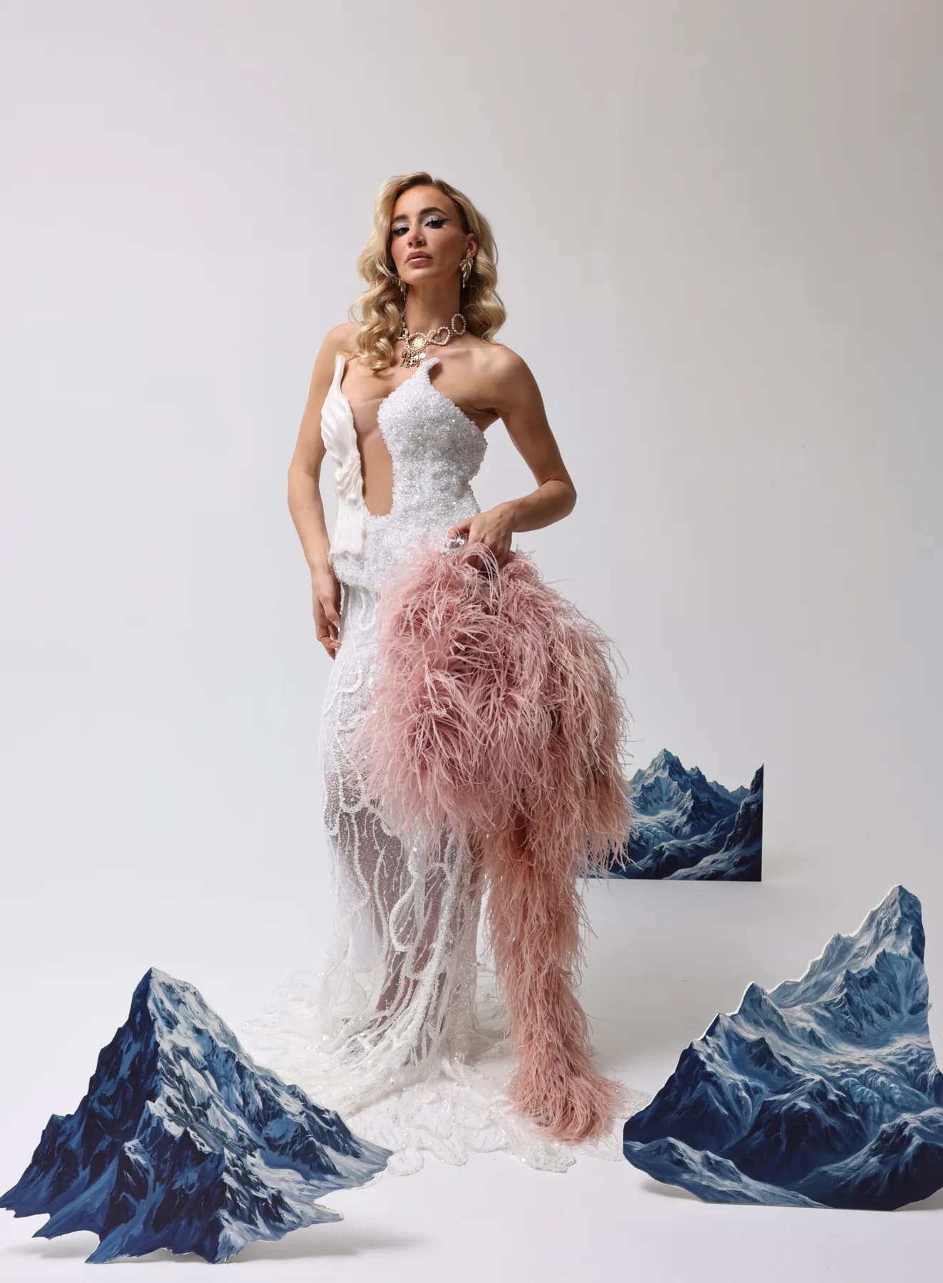 Model in a white dress holding a pink feathered piece among mountain props